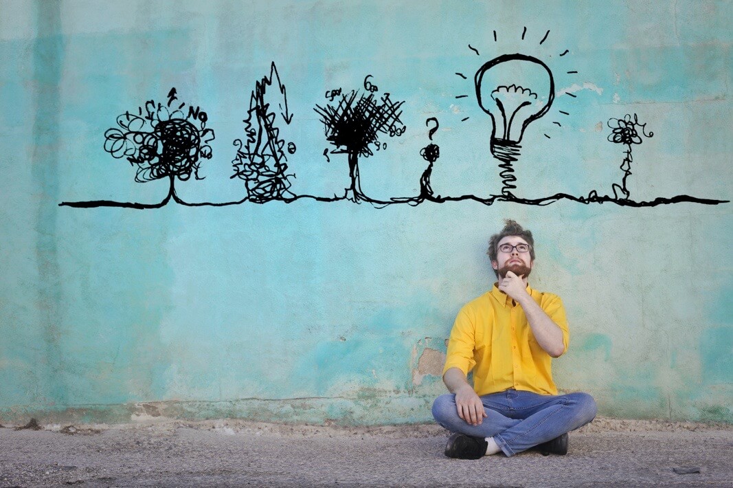 A man sits by a wall thinking with a lightbulb drawn above him.
