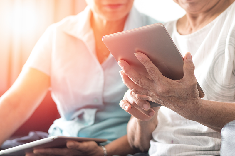 Two elderly women sit next to each other and use tablets.
