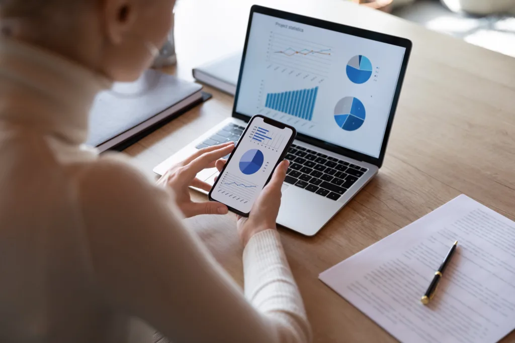 A woman holds a phone near her laptop. Both screens show blue graphs and other visual content marketing