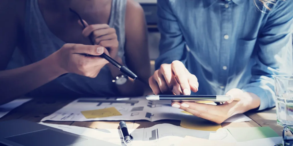 Two people sit at a desk and point at a tablet. The desk is covered in papers.