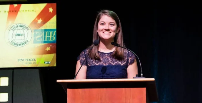 Mackenzie Bender stands behind a podium on stage wearing a navy blue blouse.