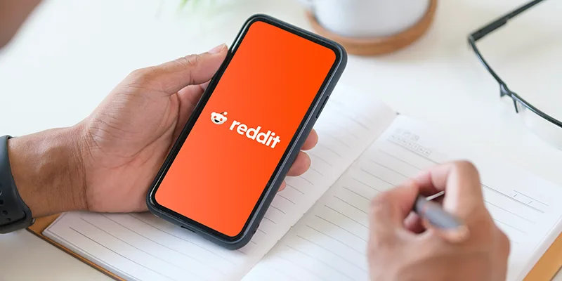 Hands holding a phone with the Reddit app open while taking notes, illustrating the concept of using Reddit for marketing