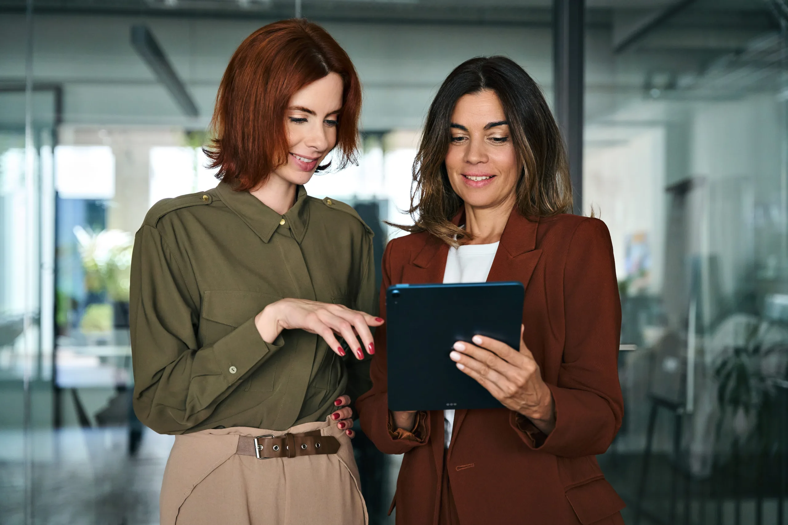 Two professional women in a modern office environment collaborating and reviewing digital marketing analytics for SEO services on a tablet.
