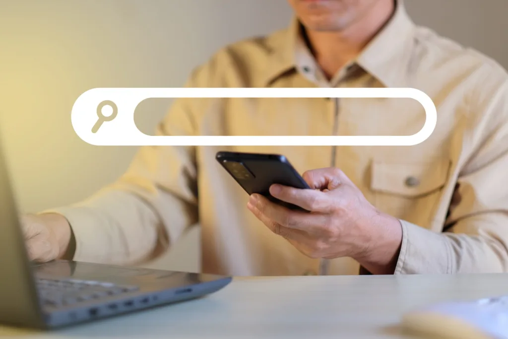 A man using his phone and laptop to with a search bar superimposed over the image.