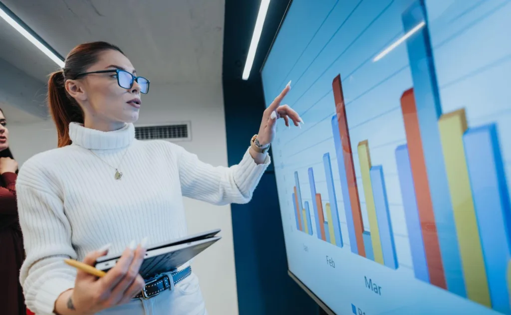 A businesswoman holding a laptop and pointing to a large digital screen displaying bar charts, demonstrating the results of various content marketing analytics tools.