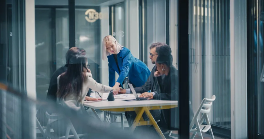 A diverse corporate team collaborating in a glass-walled meeting room, leaning over a desk to review printed content analytics and data reports.