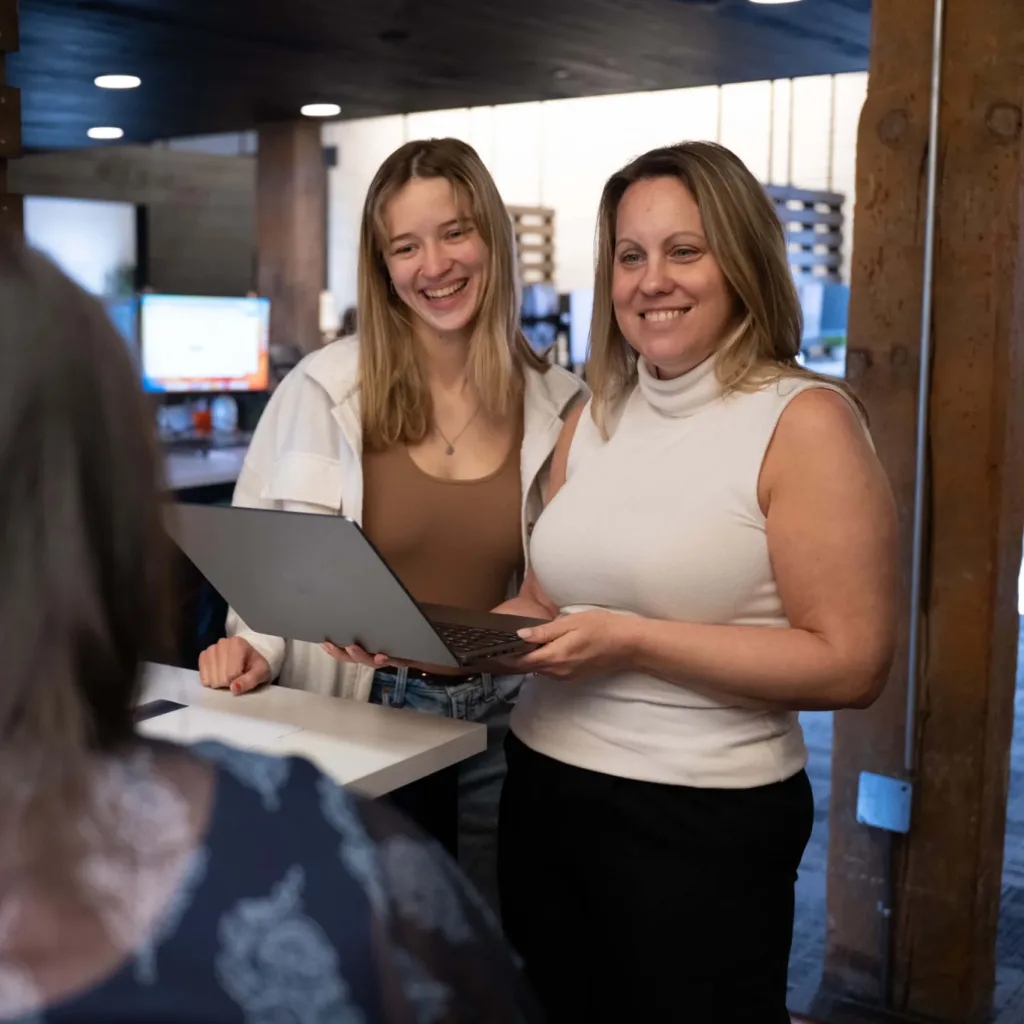 Two female employees of a restaurant digital marketing agency stand at a desk and smile. One of them holds a laptop.